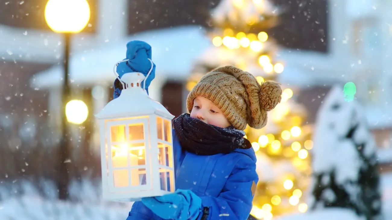 Child in a knit hat holding a lantern on a snowy small-town street