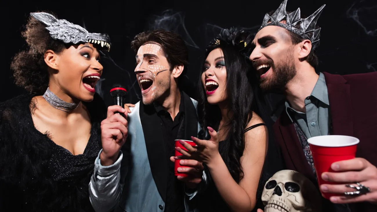 Joyful multiethnic friends in spooky costumes singing karaoke on halloween party on black
