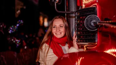 Smiling woman in a red scarf beside an illuminated truck at a night parade