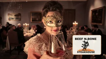 Guest in a gold masquerade mask raising a glass at a candlelit dinner, with the Beef N Bone Steakhouse logo