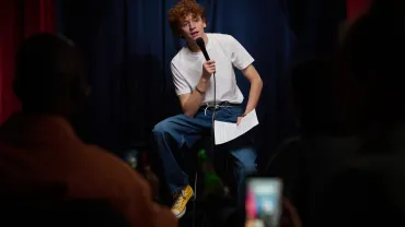 Person performing stand-up comedy on stage holding microphone and script in hand. Audience members visible with phones c