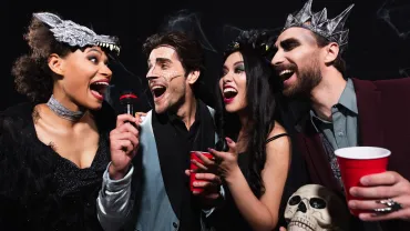 Joyful multiethnic friends in spooky costumes singing karaoke on halloween party on black