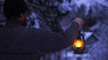 A man with a kerosene lamp in his hand walks through the snowy forest at night to his house in the village