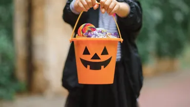 Little cute girl in witch costume holding jack-o-lantern pumpkin bucket with candies and sweets. Kid trick or treating i