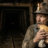 An 1890s Cornish miner in a felt hat with a candle stuck on with clay, holding a pasty, hears a tommyknocker: red eyes glow in the dark mine tunnel behind him (AI-generated illustration)